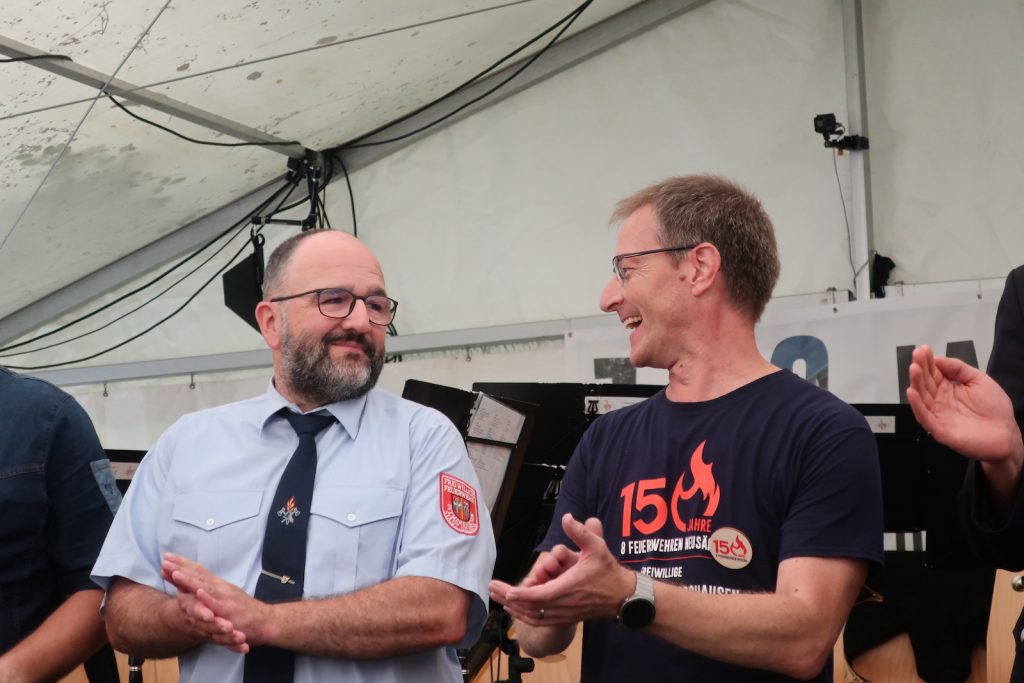 Die beiden Geehrten stehen vor der Bühne und blicken sich fröhlich an, während sie applaudieren.